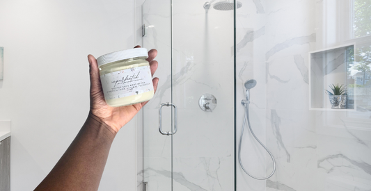 Hand holding shea butter cream in front of glass shower